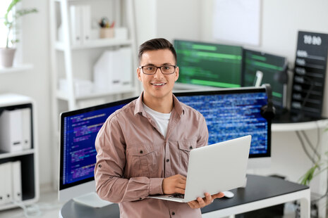 Junger Mann am Laptop sitzend, hinter sich weitere große Computerbildschirme