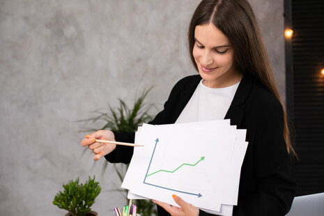 Junge Frau mit langen Haaren hält in der linken Hand mehrere Papierbögen, auf dem ersten ist ein Diagramm zu sehen. In der linken Hand hält die Frau einen Stift und zeigt auf das Diagramm.